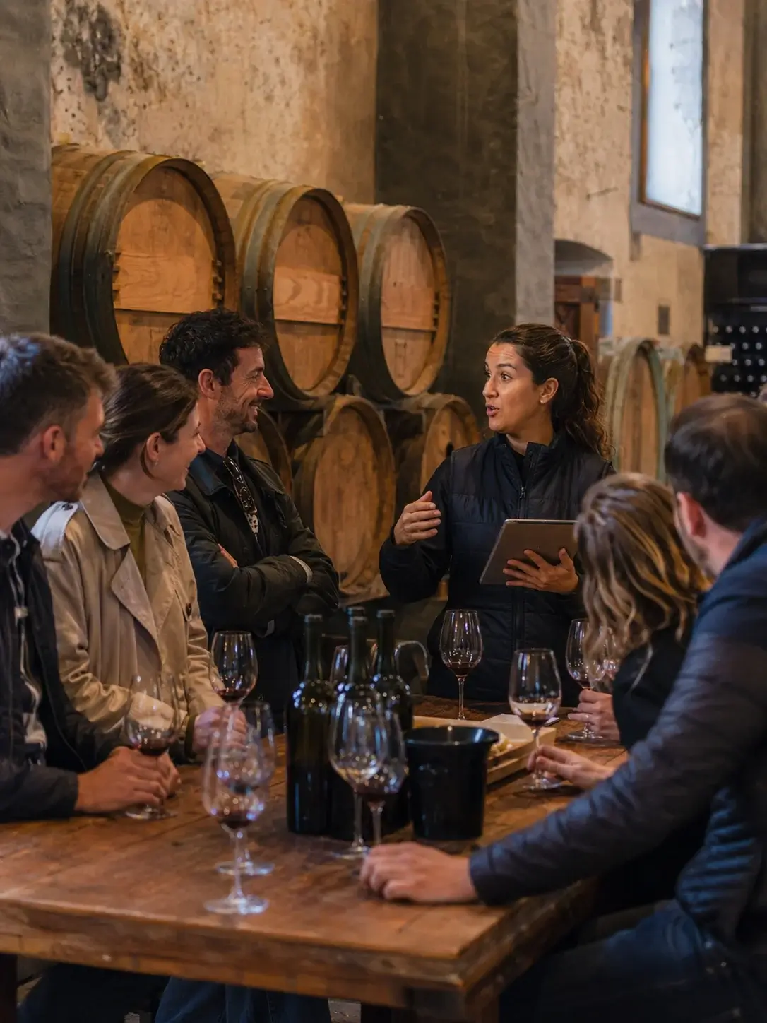 A group of visitors enjoying a guided wine tasting at a winery, with glasses and wine barrels.